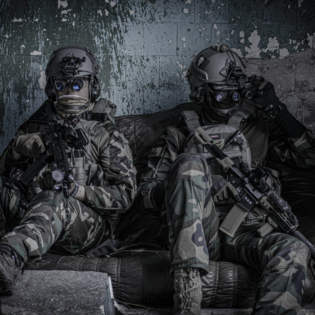 Two soldiers in tactical gear sitting on a worn couch, with rifles and focused expressions.