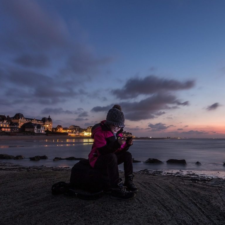 Person in a pink jacket sitting by the beach at sunset, with city lights in the background.