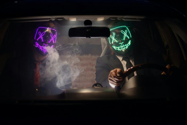 Two individuals in a car wearing glowing masks, with smoke inside.