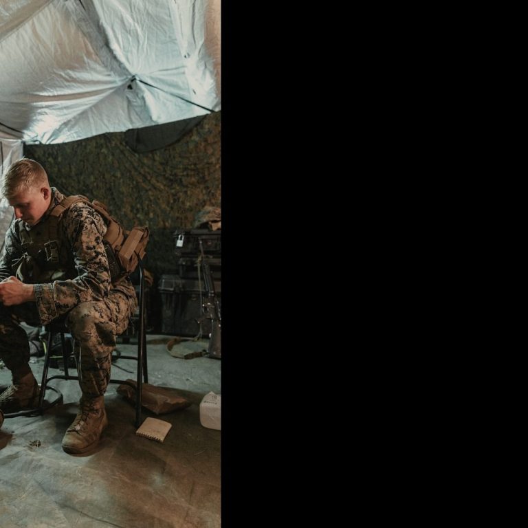 Soldier in camouflage gear sits at a desk in a military tent, focused on a device.