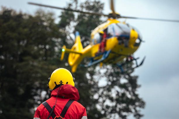 Crisis Response Rescue worker in a yellow helmet watches a helicopter approaching a forested area.