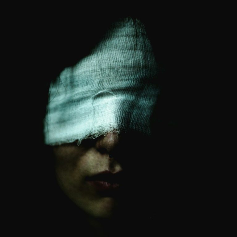 A face partially obscured by a dark cloth, with shadows creating a dramatic effect.