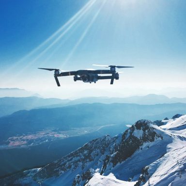 A drone flying over snow-covered mountains and a valley under a bright sky.