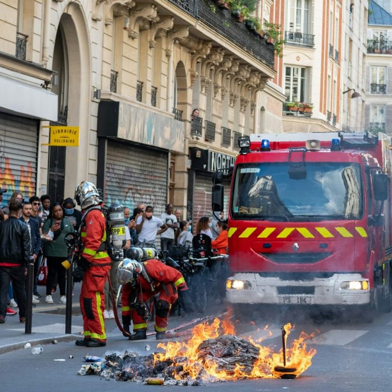 Firefighters extinguish a blaze in the street near a fire truck and a crowd of onlookers.