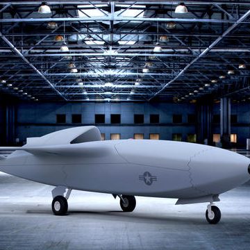 A sleek, white unmanned aircraft in a spacious hangar with industrial lighting.