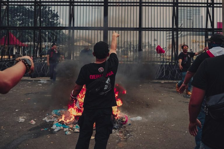 Protesters gather around a fire, raising their fists amidst smoke and chaos.