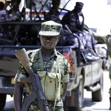 Soldier in tactical gear stands in front of military truck with armed personnel.