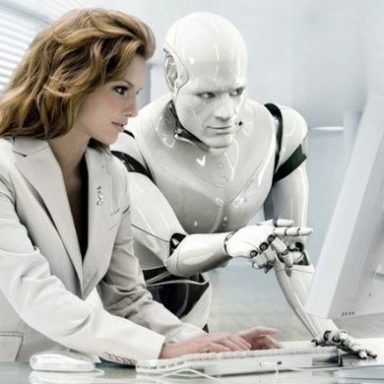 A woman in a suit working at a computer with a humanoid robot beside her.