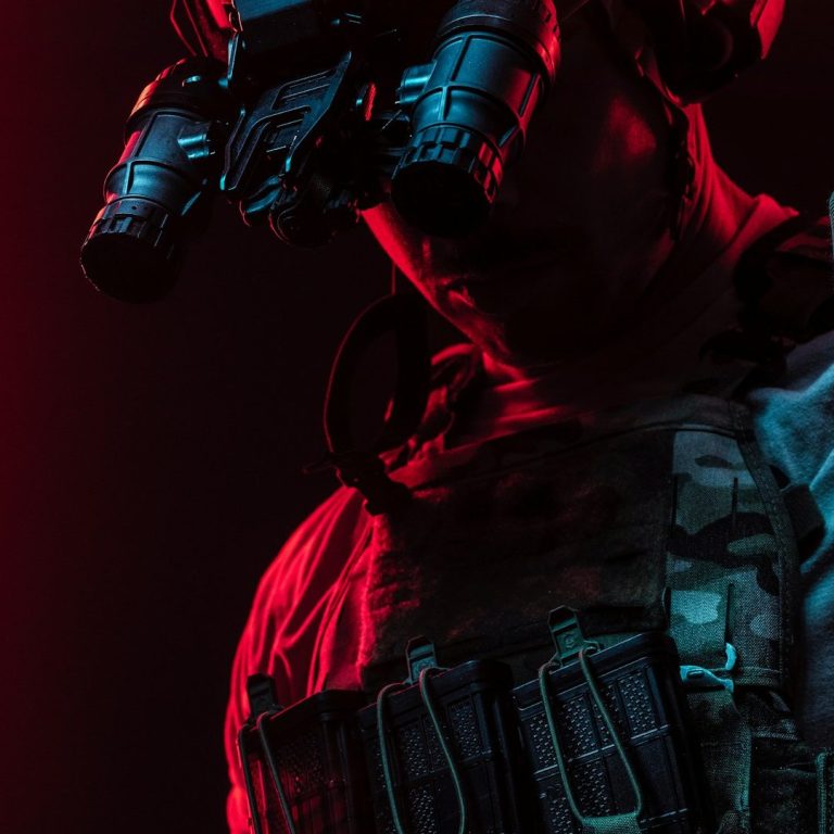 Soldier in tactical gear with night vision goggles against a dark, red-lit background.