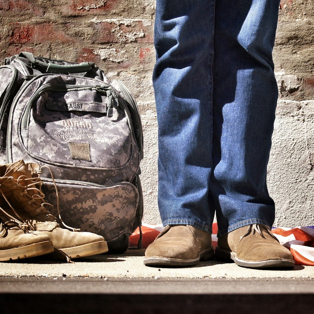 A pair of brown shoes, jeans, and a camo backpack on a rustic surface.
