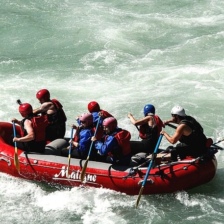 Six people in a red raft navigate turbulent water, wearing helmets and paddling.